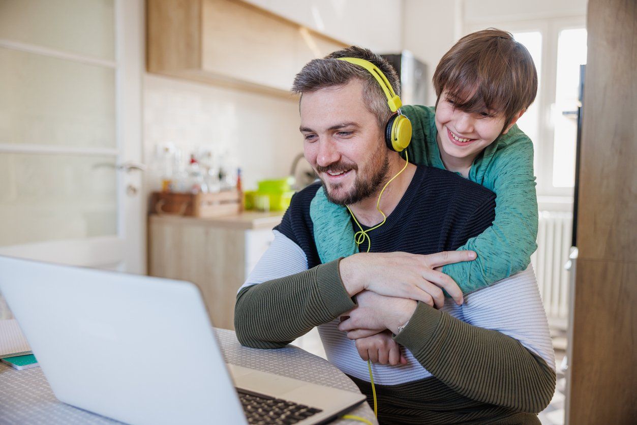 a photo of a dad on his computer wearing headphones and his son on his back.