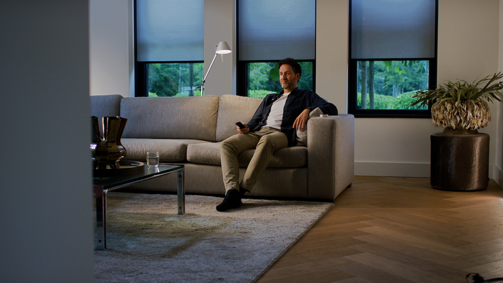 a photo of a man sitting on a couch in a room with Eve MotionBlinds on the windows