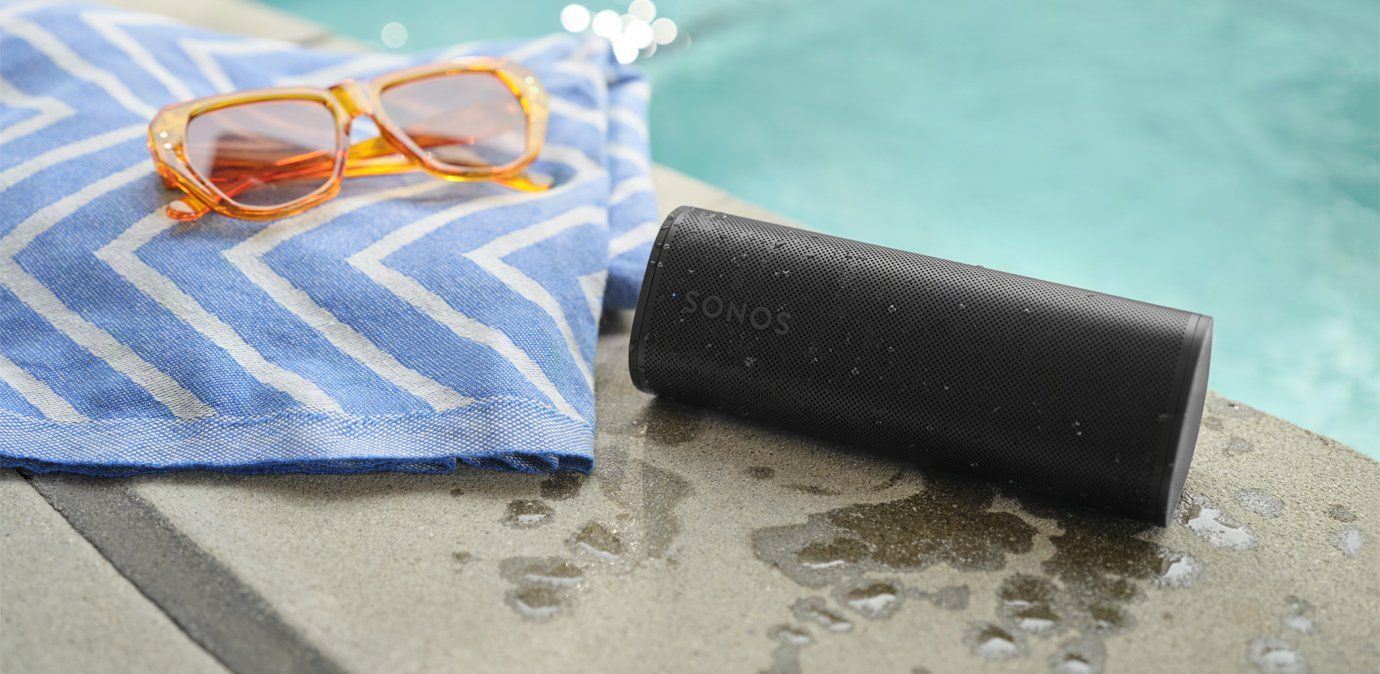 a photo of Sonos Roam 2 Portable Wireless Speaker on a pool deck next to a towel with sunglasses on top.